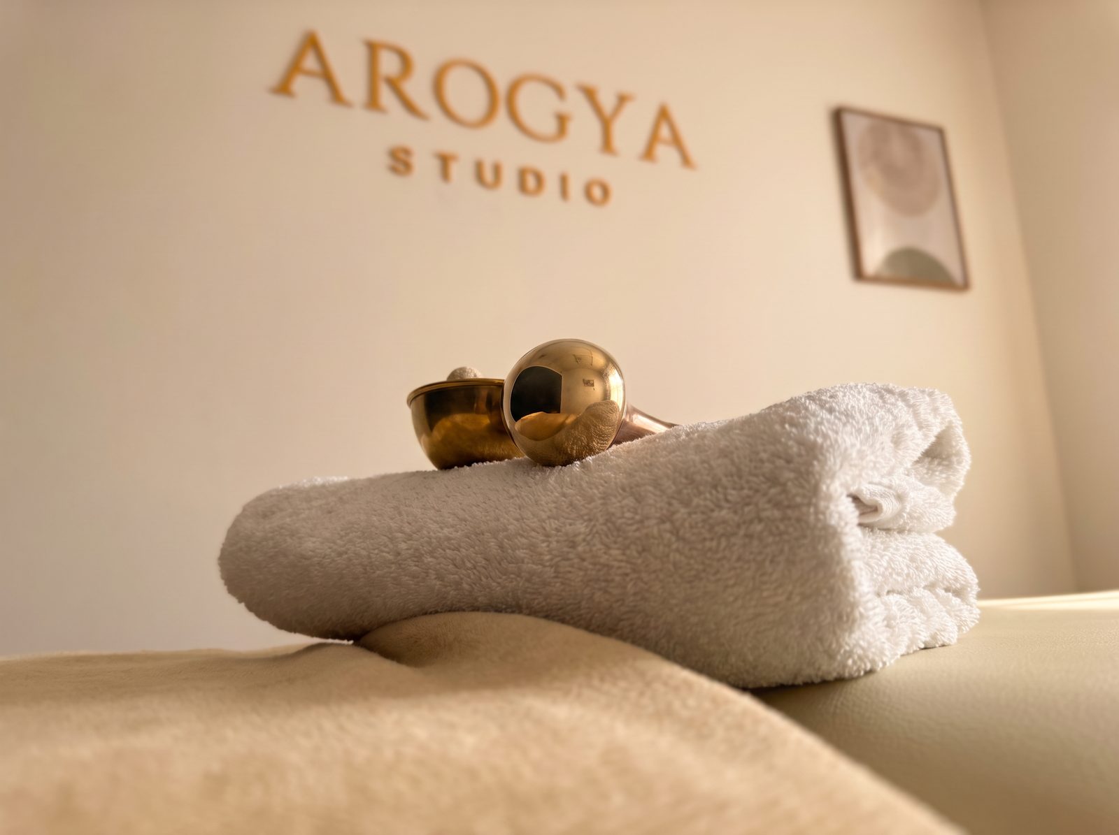 Bronze Kansa massage wands on a folded towel at Arogya Studio, with the studio sign behind